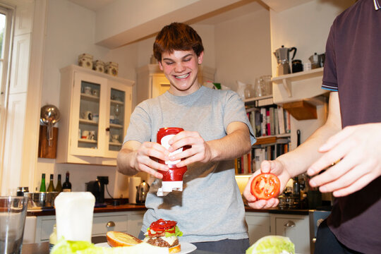 Happy Teenage Boy Squeezing Ketchup Onto Hamburger In Kitchen