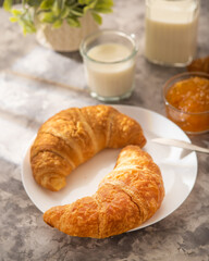 Croissants on a white earthenware plate. In the background, orange jam, milk in a glass jug and glass. White background. High angle view. There are no people in the photo.