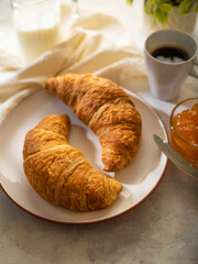 Two croissants on a white earthenware plate. An amber orange jam in a glass bowl and a cup of aromatic black coffee sit on a table covered with a white silk tablecloth. High angle view.