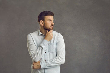 Portrait of serious bearded man rubbing chin deep in thought looking to side standing on grey ad text background. Handsome entrepreneur or student thinking over interesting idea or wise business quote