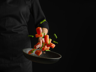 A cook in a black uniform holds a frying pan with pieces of red fish and vegetables in a levitation state. Focusing on the foreground. Place for your lettering.