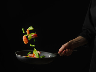 Pieces of trout with vegetables and herbs in a frying pan in a state of levitation. The black background and the bright colors of the ingredients create a beautiful contrast.