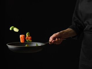 The process of cooking red fish with vegetables in a frying pan. Fish and vegetables in a state of levitation. Black background. Focusing on the foreground.