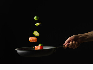 Pieces of red fish and vegetables in a state of levitation. The chef's hand holds a frying pan with fish and vegetables. Black background. Minimalism. Wallpaper. Texture. Background.