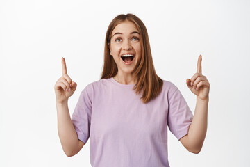 Portrait of excited blond young woman, caucasian girl pointing and looking up with amazed, happy...