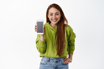 Cute teenage redhead girl, show mobile phone app and smiling happy, introduce smartphone...
