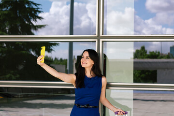 Smiling businesswoman making selfie photo on yellow smartphone. Wearing blue dress. Standing on...