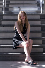 Vertical full-length view of beautiful teenaged girl with long dirty blond hair dressed in a short black dress sitting in stairs in dappled light