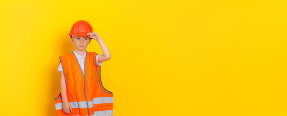 Portrait of a young (child) handsome worker or engineer wearing a helmet and orange reflective vest on a yellow background. The boy holds the helmet with his hand