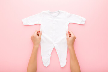 Young adult mother hands holding white new baby pajama with long arms and legs on pink table background. Pastel color. Closeup. Point of view shot. Empty place for text on apparel. Top down view.