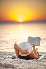 Fototapeta na wymiar Young woman reading a book at the beach while the sun rise. She's wearing a orange dress and white hat