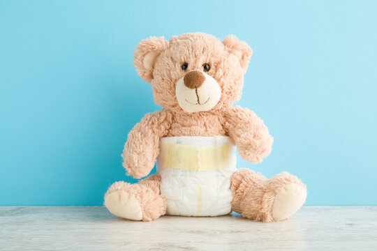 Brown teddy bear in white soft baby diaper on table at light blue wall background. Pastel color. Closeup. Front view.