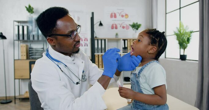 Handsome confident professional black-skinned male doctor examining small girl's throat with spatula in his medical office in modern treatment center