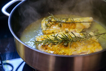 Fish fillet being fried in frying pan with rosemary and capers.