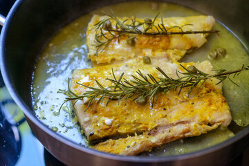 Fish fillet being fried in frying pan with rosemary and capers.
