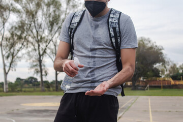 Detail view of a young man wearing a face mask applying alcohol spray to his hands in an abandoned field.