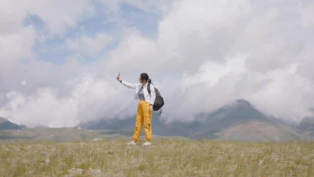 Tourist takes selfie on smartphone camera in mountains. Young woman traveler with backpack shoots video. Travel blogger on hike in mountains against backdrop of clouds