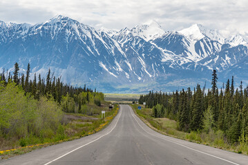 Alaska Highway driving into Haines Junction town in spring time with epic, huge mountains in far distance with amazing scenic drive ahead. Tourists, tourism shot for camping, RV, road trip. 