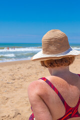 An adult woman in a swimsuit and a hat sits on the seashore on a sunny day, rear view. Summer vacation, travel and tourism