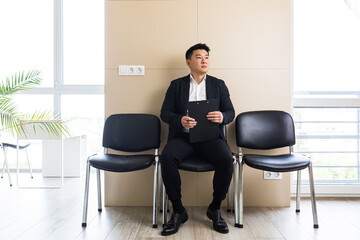 Young successful Asian man, waiting for a job interview, in the waiting room of the office center, sitting on a chair near the reception of the office center