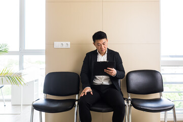 Young successful Asian man, waiting for a job interview, in the waiting room of the office center, sitting on a chair near the reception of the office center