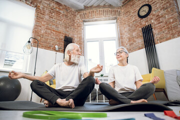 Two aged people in activewear sitting in lotus position on yoga mat and meditating with closed eyes. Domestic exercises for mental health. Aged family training at home.