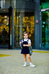 a beautiful Caucasian girl, a schoolgirl, is standing near the school, with a backpack and holding a textbook in her hands