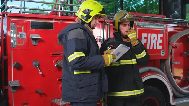 One fireman use tablet and discuss with his co-worker who contact other people in teamwork by using walkie talkie. They stand in front of fire truck during plan to work firefighting.