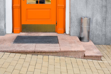 granite threshold with foot mat near orange wooden front door with iron trash can near building...
