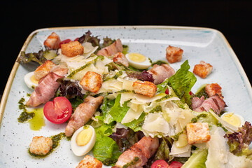 Caesar salad with herbs chicken bacon eggs and tomatoes on a square light plate, on a dark background