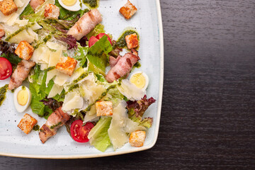 Caesar salad with herbs chicken bacon eggs and tomatoes on a square light plate, on a dark background