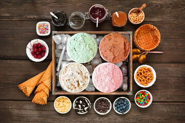 Summer ice cream buffet with a variety of flavors and sweet toppings. Overhead view table scene on a dark wood background.