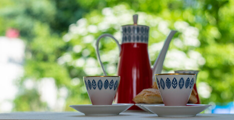 Red teapot with blue pattern teapot and two white cups on a wooden table facing the street. Flowers in the background. Morning tea, breakfast.