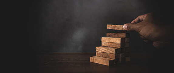 Close-up hand is placing wood block tower stacked in stair step with caution to prevent collapse or...