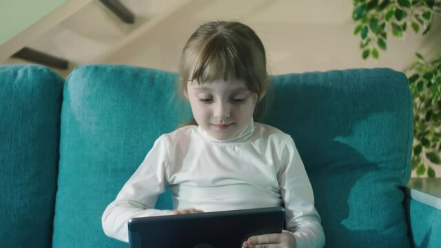 Preschooler Girl Playing Games Using Digital Tablet Lying On Sofa Alone. Curious Little Kid Girl Hold Pad Computer Watching Video At Home Children Education Or Tech Internet Addiction Concept