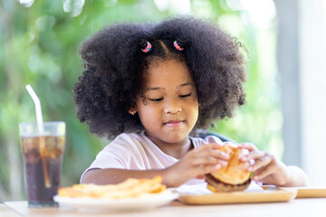 A little curly-haired African-American girl sits on the table with a knife and eats delicious...