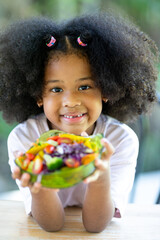 Little African American curly haired girl sits and eats vegetable. delicious salad on the table Fun, cheerful. Appetite. Childhood and eating concepts.