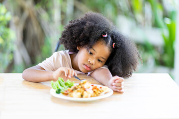 A young, curly haired African American girl sits looking at food. Childhood concepts and healthy eating.