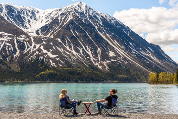 Couple sitting outdoors in mountains of Kluane National Park, Yukon Territory, northern Canada with picnic, red table between and camping, lounging chairs. Stunning blue sky day. 