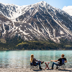 Man and woman couple sitting beside a lake in scenic mountain view setting, scene in Kluane National Park  with hige snow capped mountain peaks in distance. 