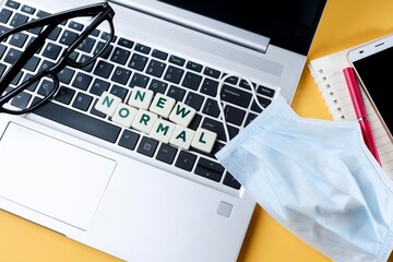 Lettering on New Normal on the laptop keyboard with a face mask, eyeglasses, and smartphone and pen on the notebook