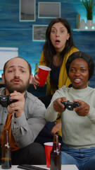 Woman with dark skin losing videogames against friend man while playing for gaming competition using joystick. Multi-ethnic friends drinking beer having fun together sitting on couch late at night
