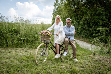 blonde woman with white dress and her boyfriend or husband posing with bicycle with beautifully decorated flower basket in nature and are happy and in love, nature concept, flowers, delivery service