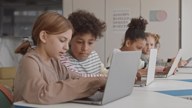 Medium shot of small group of smart multiethnic schoolchildren studying on laptops in modern IT class