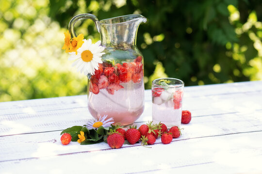 Homemade strawberry mojito lemonade in a glass jug on a white wooden rustic table. Strawberries, mint sprigs and flowers. Summer ice cold drink.