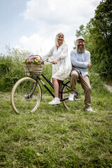 blonde woman with white dress and her boyfriend or husband posing with bicycle with beautifully decorated flower basket in nature and are happy and in love, nature concept, flowers, delivery service