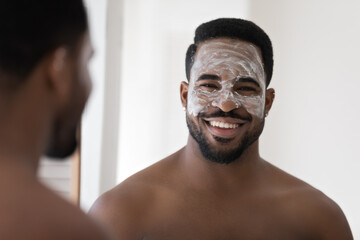 Happy attractive African guy with white cleansing natural scrub, cosmetic mask on face looking at...