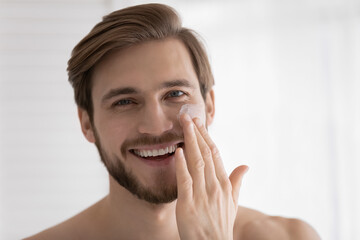 Head shot of young attractive guy applying moisturizer, sunscreen on face, applying anti age collagen cream, mask on under eye skin, preventing sun burn, wrinkles, dryness. Close up portrait