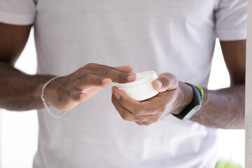 Hands of mixed raced Black man in white t-shirt, taking moisturizing collagen cream, lotion, mask,...