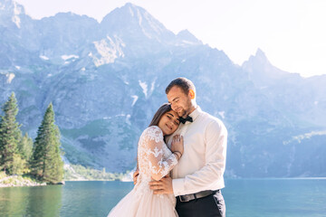 A young couple of newlyweds, the bride and groom are walking along the rocky shore of a mountain lake against the backdrop of beautiful mountains and blue sky. The bride gently hugs the man